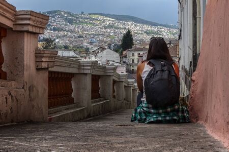 A woman sitting on old stairs of the old town of Quito, Ecuador. She is with her back turned and a beautiful mount landscape of the village can be seen behind.の写真素材