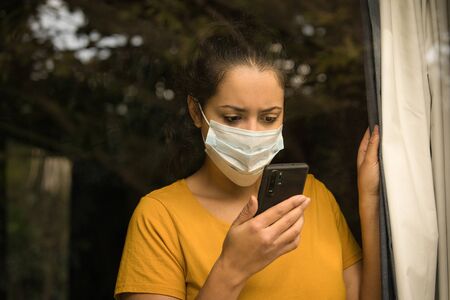 Latin woman wearing a face mask looking at her smartphone, self isolated at home. COVID-19 and Quarantine concept.の写真素材