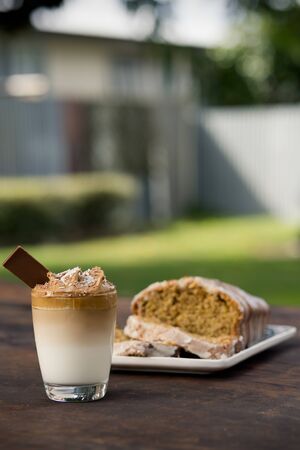 Cup of Dalgona coffee served on a glass with a bar of chocolate and a wholemeal cake behind. Outdoor backgroundの写真素材