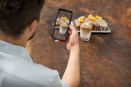 Man taking a photo with mobile at a glass of Dalgona coffee and an orange cake on a wooden...