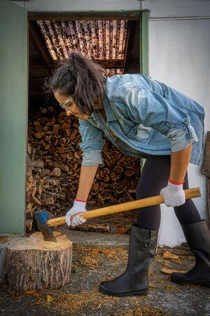 Young brunette woman chopping wood with an ax at home. She is wearing gum boots, protective glasses and gloves. Vertical photographyの写真素材