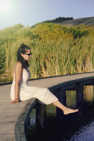 Young brunette woman wearing dress sitting on a wooden walkway and relaxing on barefoot. Vertical photography. Inspirational conceptの写真素材