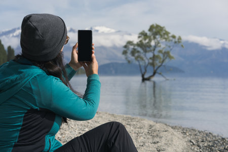 Woman taking picture with smartphone at iconic Wanaka Tree. New Zealand famous placeの写真素材