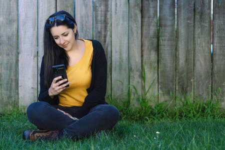 Brunette woman sitting on the grass while looking at her cellphone. Social Medial Conceptの写真素材