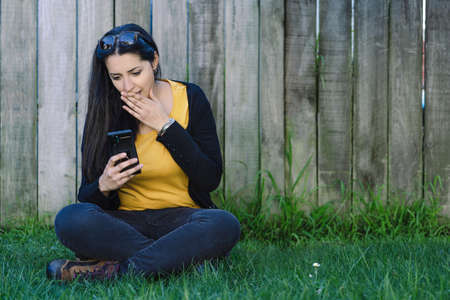 Young hispanic woman sitting on the grass and looking at her mobile with a surprise face. Technological conceptの写真素材