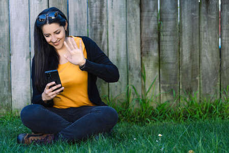 Latin woman sitting on the grass while having a video call with her cellphone. Technological concept.の写真素材