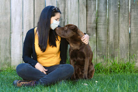 Woman wearing a face mask is sitting on the grass with his dog at the park. Social distancing and conceptの写真素材