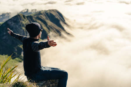Young man sitting at the peak of a mountain and opening his arms on a foggy morning. Travel conceptの写真素材