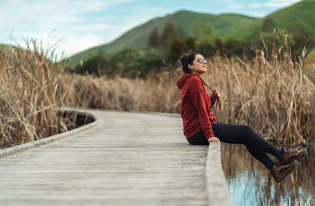 Hispanic woman wearing casual clothes sitting on a wooden walkway surrounded by a lake. Peka Peka Wetlands, Hawke's Bay, New Zealand.の写真素材
