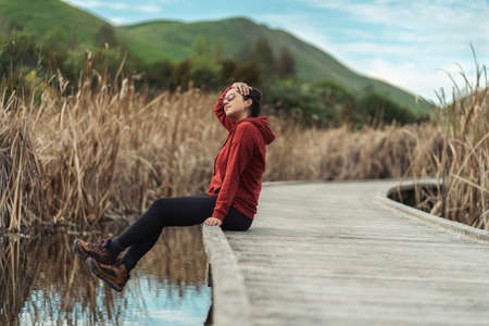 Young brunette woman relaxing at the edge of a wooden walkway on a lake. Hillside and wetlands. Inspirational conceptの写真素材