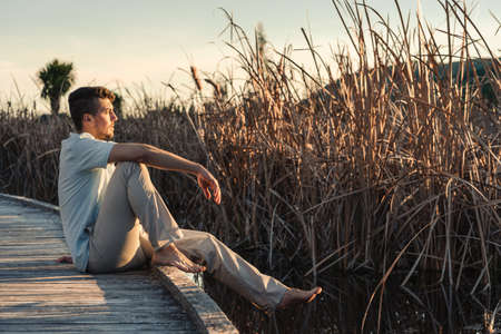 Young man posing in profile on bare feet sitting on wooden walkway on lake shore. Portrait photography at sunsetの写真素材