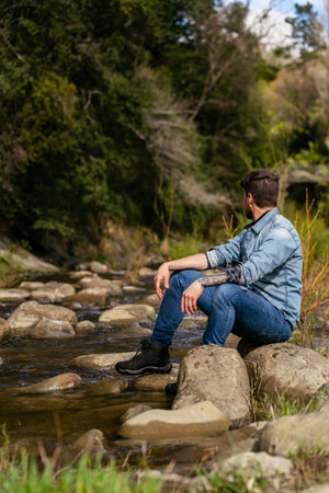 Young man admiring the natural environment that surrounds him while sitting at edge of riverの写真素材