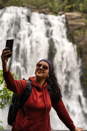 Hispanic woman taking selfie with mobile at lookout of waterfall. Travelers concept. Vertical photographyの写真素材