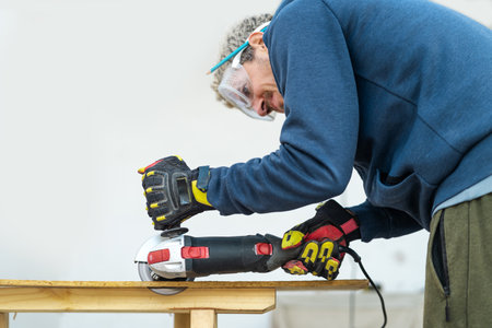 Man with electric saw to cut wooden piece at home. He is wearing protective glasses and gloves. close-upの写真素材