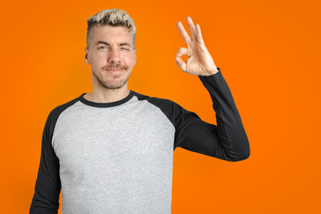 Caucasian man doing OK gesture with hands and winking. Casual wear. Isolated orange backgroundの写真素材