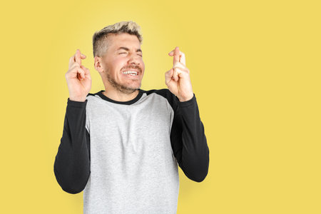 Excited blond hair man with fingers crossed and closing his eyes posing on yellow background. Copy space on right sideの写真素材