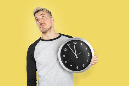 Tired and bored Caucasian man looking up like exhausted of waiting while holding wall clock. Standing on isolated yellow backgroundの写真素材