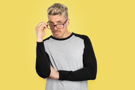 Blond hair man in casual clothing adjusting eyeglasses and looking curious at camera. Portrait photo on studio shot with yellow backgroundの写真素材