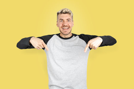 Friendly Caucasian man in casual clothing doing smiling at camera and pointing fingers down for advertisement offer copy space. Isolated studio shot on yellow backgroundの写真素材