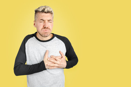 Young adult blond hair man standing and having a heart attack while holding his chest with both hands. Isolated studio shot on yellow background.の写真素材