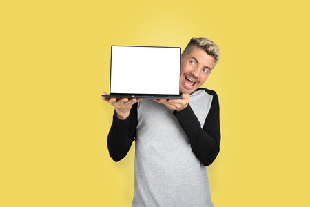 Caucasian happy man in casual clothing showing open laptop with white screen for copy space standing on yellow background. Advertisement sale concept studio shotの写真素材
