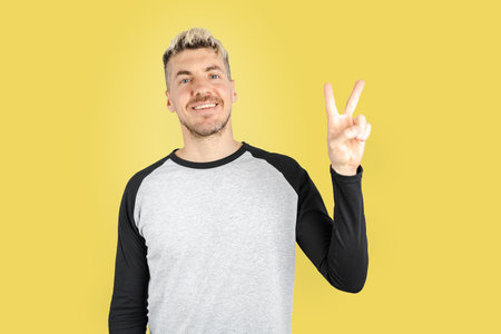 Caucasian young adult man doing victory sign with finger and smiling at camera. Isolated studio shot on yellow backgroundの写真素材