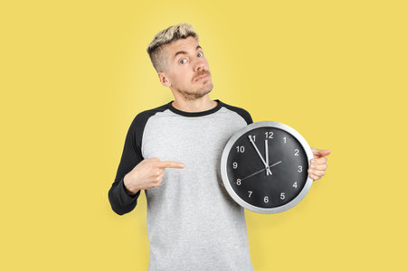 Caucasian man in casual clothing showing time on wall clock and pointing with finger with gesture of concern, Isolated yellow backgroundの写真素材
