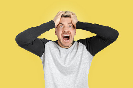 Caucasian angry man in casual clothing screaming out of control while holding his head in desperation. Isolated yellow backgroundの写真素材