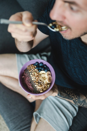 Man sitting on the couch while eating muesli and smoothie bowl with fruit and nuts for breakfast. Portrait photography. Selective focus. Healthy lifestyle conceptの写真素材