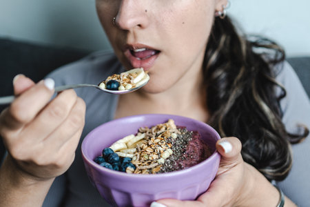 Young Hispanic woman eating muesli bowl with smoothie, nuts and fruit at home. Close-up shot. Unrecognizable person. selective focusの写真素材