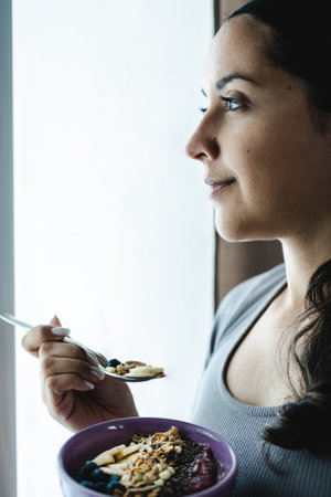 Hispanic Latin woman standing in profile next to window in the morning while eating delicious bowl of muesli with smoothie, fruits and nuts. Well-being and nutrition concept. portrait photoの写真素材