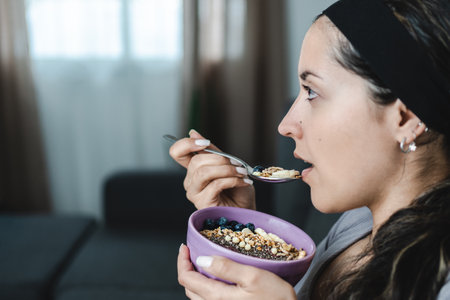 Young Latina sitting in profile eating muesli bowl for breakfast with fruits and nuts. Close up caption. Healthy lifestyle conceptの写真素材