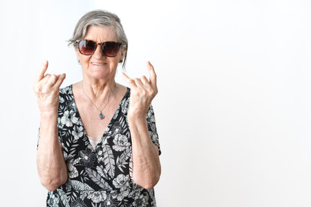 Senior woman wearing sunglasses smiling and doing rock symbol with hands up with arthritis. Isolated white background studio shotの写真素材