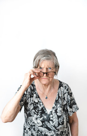 Skeptical woman looking at camera while peering over her eye glasses. White isolated background. Vertical photography with copy spaceの写真素材