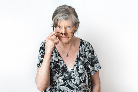 Portrait of blue-eyed angry senior woman peering over her eyeglasses on white background studio shot.の写真素材