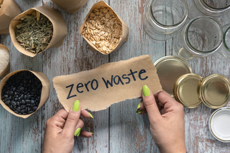Woman hand holding paper that says zero waste on wooden table with bulk food and recycled glass...