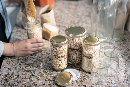 Table in the kitchen with bulk food in recycled jars and woman behind taking groceries from paper bags. Close up shot. Zero waste at home concept. selective focusの写真素材