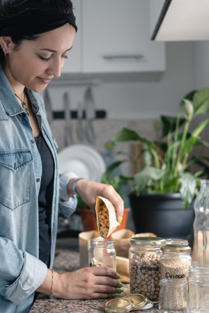 Latin American woman putting bulk food in recycled glass jars in the kitchen. Plastic free and zero...