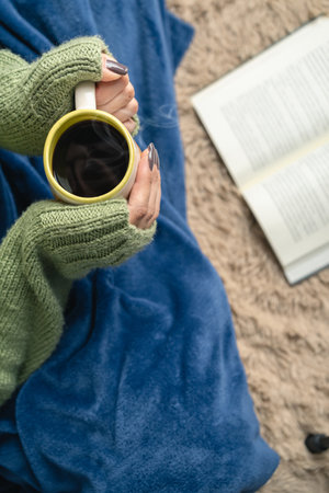 Top view of woman hand wearing cozy wool sweater holding mug of coffee while reading a book on the floor of living room. Vertical photography. Copy space for textの写真素材