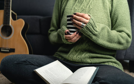 Unrecognizable woman with cozy knit sweater holding cup of coffee and reading book in the living room carpet. Call for cozy winter conceptの写真素材