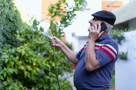 Senior old man talking on the mobile phone while watering his garden. Enjoyment in the old age conceptの写真素材