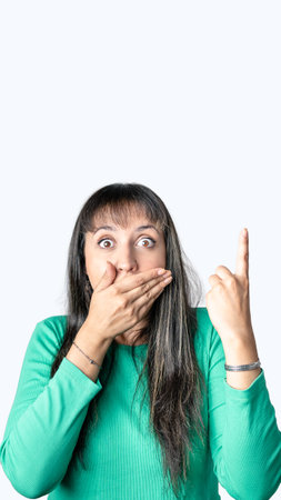 Surprised young Latin woman covering her mouth with one hand and pointing above with the other one. Isolated white background with copy space for advertisementの写真素材