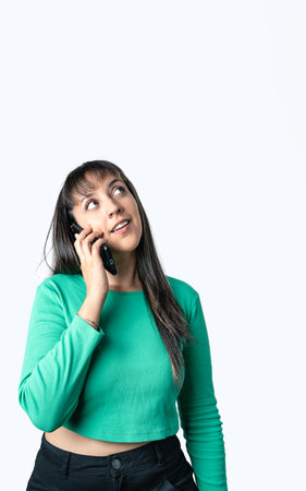 Latin woman dressed with casual clothes is talking on the phone and looking up on a white background. Vertical shot with copy spaceの写真素材