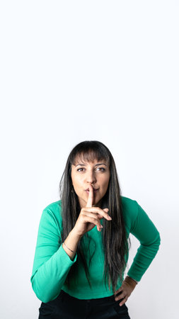 Brown hair woman with Hispanic features making silence gesture with her finger on lips over isolated white background,. Vertical photography with copy space aboveの写真素材