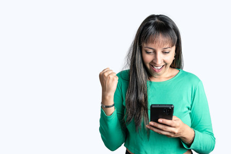 Happy Hispanic woman dressed in casual clothes looking at her smart phone screen and celebrating on white isolated background. Copy space on left side for advertisementの写真素材