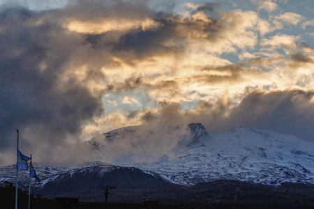 Snow-covered mountain peak of Copahue volcano with dramatic clouds with Argentina flags visible in the foreground. Cavioahue, Neuquen, Argentinaの写真素材