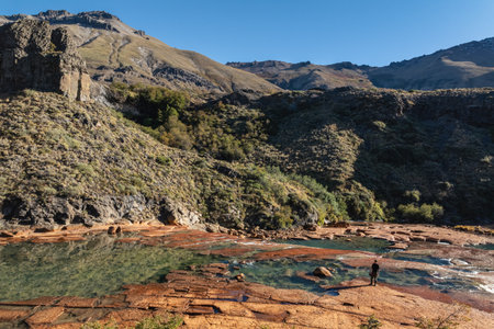 Man standing by the rocky, shallow Agrio River with mountains rising in the background under a clear sky. Neuquen, Argentinaの写真素材