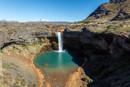 Salto del Agrio waterfall plunges into a turquoise pool surrounded by rocky terrain, with snow-capped Copahue volcano visible in the distance. Neuquen, Argentinaの写真素材