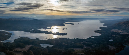 Aerial view of a Lake Alumine reflecting the sunlight and surrounded by forested hills and distant mountains under a dramatic cloudy sky. Villa Pehuenia, Neuquen, Patagonia, Argentinaの写真素材