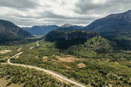 Winding road travels through a lush green valley surrounded by araucaria trees growing on rocky mountainside under cloudy sky. Neuquen, Patagonia, Argentinaの写真素材